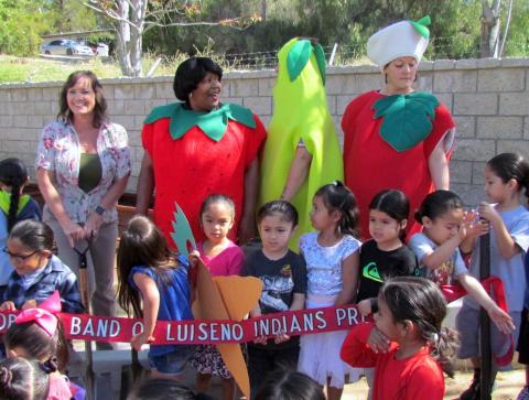 Mary Rose Morreo, center, was given the honor of cutting the red ribbon to officially open the Soboba Tribal Preschool garden on April 17. Her mom, Anita Morillo, launched the idea of farm-to-table food when she was a chef at the preschool’s kitchen. Standing behind Mary Rose to lend a hand are, from left, preschool director Dianne King, Health Education Assistant II Hosea Jones (strawberry), preschool teacher Melissa Arviso (pear) and classroom aide Amber Lopez (apple) Mary Rose Morreo, center, was given the honor of cutting the red ribbon to officially open the Soboba Tribal Preschool garden on April 17. Her mom, Anita Morillo, launched the idea of farm-to-table food when she was a chef at the preschool’s kitchen. Standing behind Mary Rose to lend a hand are, from left, preschool director Dianne King, Health Education Assistant II Hosea Jones (strawberry), preschool teacher Melissa Arviso (pear) and classroom aide Amber Lopez (apple)